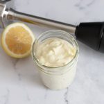 Szklany słoik z gęstym kremowym majonezem obok ręcznego blendera i składników